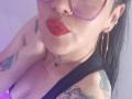 LilithRossi with red lipstick, hoop earrings, nose ring, glasses, tattoos, black tank top, showing her tits, hand on neck, purple light, black hair.