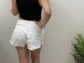 Tilliana's ass in white, frayed denim shorts, black tank top, hand on hip, standing with a green plant on the right. Her skin is light, and she's facing away.