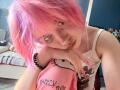 KrystalBerkovitch with pink hair, white tank top, and black bracelet, leaning on a Hello Kitty pillow, gazing at camera with long pink nails, in a sunlit bedroom.