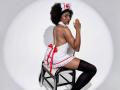 RoldanStefy in a nurse outfit with a red cross hat, white dress, red ribbon, lace trim, black thigh-highs, sitting on a chair, looking over shoulder, afro hair.
