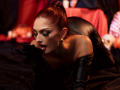 JessieDiaz with red hair in a bun, black leather gloves, and dark makeup, licking black latex, lying on black leather, showing off her tits and ass.