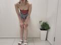 JoannaSia with bare shoulders, red strap top, denim shorts, white sandals, bent over, hands on knees, puffy legs, small tits, brown hair, necklace, white wall, potted plant.