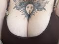 DiLaria's large tits with a sun-faced tattoo between them, featuring flowers and leaves, wearing a brown bra with gold clasps, showing her pale, slightly textured skin.