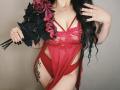 Niferttiti wearing red lace lingerie with cutouts, holding black roses, long black hair, black nails, tattoo on right thigh, gold necklace, red panties, ring on left hand.