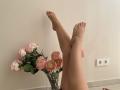 MysteryLaddy's legs are crossed at the ankles, with pink-painted toenails, next to a glass vase of pink roses on a white floor. Two electrical outlets are visible.