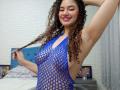 SanetPerlas with curly brown hair, smiling, wearing a blue fishnet top that exposes her tits and pussy, raising her right arm. Bedroom setting, white brick wall.