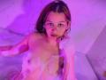 Elodie Gardner, nuda, in una vasca da bagno illuminata di viola, con bolle schiumose che le coprono i seni e la vulva, indossa degli occhiali, con i capelli castani bagnati e un'espressione sorpresa..