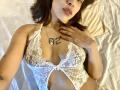 KiaraHernandezX lying on a bed, wearing a white lace bralette that reveals her small, perky tits and a tattoo on her chest. Her hand touches her collarbone.