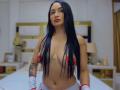 PamelaLiss with long black hair, red nipple pasties, and white gloves, standing topless, showing her tits and a tattoo on her right side, in a bedroom.