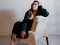 Niferttiti with wavy brown hair, wearing a black dress and green shirt, sits in a white textured chair, hand on head, looking up, legs crossed, red nails.