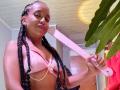 LilithAndAsmodeo with dark braids, pink bikini top, pearl necklace, holding pink dildo, smiling, light brown skin, ceiling above, green plant on right, wooden door in background.