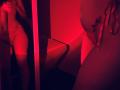 Red-lit photo showing Tisia's bare ass and Tits in lace underwear, reflection in mirror, hand on hip, tattoo on right breast, finger pointing, soft shadows, sensual atmosphere.