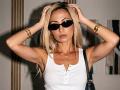 ViktoriiaLu with blonde hair, wearing black sunglasses, a white tank top, gold bracelets, and a black shoulder bag, hands in hair, looking serious.