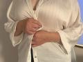 Elliance's hands are buttoning a white satin shirt, covering her tits, with her fingers slightly pulling the fabric, showing a bit of her black underwear beneath.