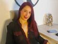 SweettEve with red hair, black sheer blouse showing her tits, red lips, sitting, smiling, desk with lantern, phone, and black chair.