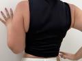 Tilliana, with tan skin, black sleeveless top, white pants, purple nail polish, and ring, stands against a wall, showing her back, with her right hand on it.