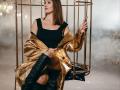 MiraMixi with brown bob, black off-shoulder top, gold metallic coat, and black knee-high boots, sitting in gold cage, holding cage bars. Boobs visible, ass slightly exposed.