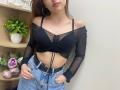 Rioria with black sheer off-shoulder top, exposing small boobs, and blue jeans with a black belt. She has long brown hair, hoop earrings, and pink lipstick.