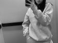 Black-and-white selfie of Olieri with pouty lips, wearing a loose gray hoodie and pants, holding a phone in front of her face. Her tits are small, covered.