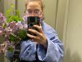 HotCaramelsTrio with curly pinkish-brown hair, glasses, and blue shirt, holding a bouquet of purple lilacs, taking a mirror selfie with a black and white phone case.