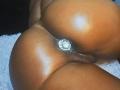 CinnamonBebe's shiny brown ass with a jeweled butt plug, glistening skin, and visible dark labia, lying on a white textured surface.