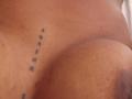 Caylin's tan, smooth, upper chest with a small black tattoo of four vertical dots, slightly to the left. Her large, brown areola is visible on the right.