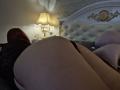 Dinastiia's ass is in the foreground, she's lying on a bed with black lace lingerie, her red hair visible, a white and gold headboard, and a lit lamp in the background.