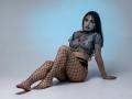 MiaHarperr with dark hair, wearing a sheer blue floral top, exposing her small tits and nipples, fishnet stockings, sitting on white floor, looking at camera with serious expression.
