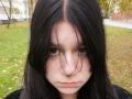 AlicaMihal has pale skin, dark straight hair, and a serious expression with her brown eyes looking forward. Her nose is slightly upturned, and she wears a black shirt.