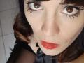OrianaLaFrancaise a les yeux marron, du rouge à lèvres, et un eye-liner foncé, avec une petite imperfection sur sa joue droite. Elle porte du noir avec une touche de tissu transparent..