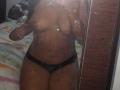 BonnieBodn's dark-skinned, topless body with small tits and black panties, reflected in a dirty mirror. She's holding her phone, covering her face. Her black dick is visible.