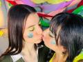 Two women kissing, ValkiriaAndDione on the left with long black hair, green eyeshadow, and a flower sticker on her cheek. ValkiriaAndDione on the right with short black hair, yellow shirt. Bright colorful background.