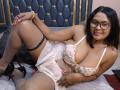 FelyLove with medium brown skin, black glasses, and straight black hair, lying on a bed in a floral lingerie dress, showing her big tits and black lace garter.