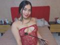 MelaniCoox with black hair, red lace bodysuit showing her tits, holding a black flogger, smiling, sitting on a bed with a red headboard. Tattoo on her right shoulder.