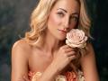 ViktoriiaLu with blonde wavy hair, holding a pink rose to her cheek, wearing a strapless dress with floral accents, smiling softly, blue eyes, fair skin.