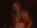 AgataRyili with red bikini, small tits, colorful nipple studs, dark red lighting, long brown hair, serious expression, wearing small gold earrings, standing against dark background.