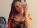 ValeryScot with curly brown hair, wearing a black lace bra that shows her tits, taking a mirror selfie with a white phone, a blue backpack in the background.