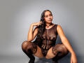 JeimyCampos squats, wearing a black lace mesh bodysuit, showing her boobs and pussy through the fabric, with lace thigh-highs, and a red pendant necklace, against a gray background.