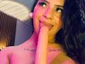 LonxEva with black curly hair, tongue out, licking a large, pink, erect dildo. She has tattoos on her neck and is topless, with pink and purple lighting.