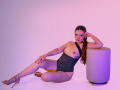 AnabelleRoss with long brown hair, sitting on the floor, wearing a black one-shoulder top, exposing her tits, and gold high heels, leaning on a white cylinder.