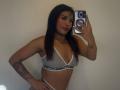 LexieBloom with dark hair, wearing a gray sports bra and white underwear, holding a phone, showing a tattoo on her left arm, and a small heart tattoo on her right.