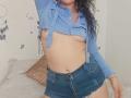 YessiSoto with wet black hair, blue shirt open to expose small bare tits, and high-waisted denim shorts, standing with arm raised, showing a slight belly.