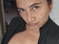 AlessiaRed with freckled face, brown eyes, and dark hair in a partial updo, wearing a black zip-up, exposing her tits. She's giving a slight, confident, smirk.