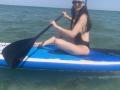 Dalaila with long brown hair, wearing a blue headband and black one-piece, paddles a blue and white stand-up paddleboard in green water, smiling with sunglasses on.