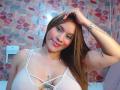BelletinaXx with brown hair, smiling, wearing a white bralette, hand on head, tattoos on arm and chest, pink lips, light skin, small tattoos visible, pink and red tile background.