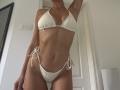 SandraSwweet trägt einen weißen, gehäkelten Bikini mit kleinen Brüsten und einem schlanken, trainierten Körper. Sie hebt einen Arm, wobei sich ein flacher Bauch und hochgebundene Seitenparts zeigen..