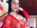 CristalZafiro, con cabello negro largo, gafas, una blusa roja de encaje que muestra sus pechos, un collar rojo, un cojín rosa, sonriendo, con la mano derecha en la mejilla, en una habitación..