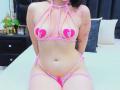 AlisonTatcher in pink strappy lingerie with heart pasties over her tits, sitting on a bed, showing her pussy through the lace, tattoo on her right arm, black hair.