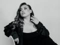 Niferttiti with wavy hair, black dress, and satin jacket, hand on neck, looking sideways, soft smile, gray background, ring on finger, necklace, subtle makeup, sexy.