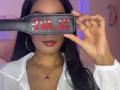 NatLewis holding a black leather paddle with "VAJ" in red letters, covering her eyes. She has dark skin, long black hair, red lipstick, white shirt, and gold necklace.